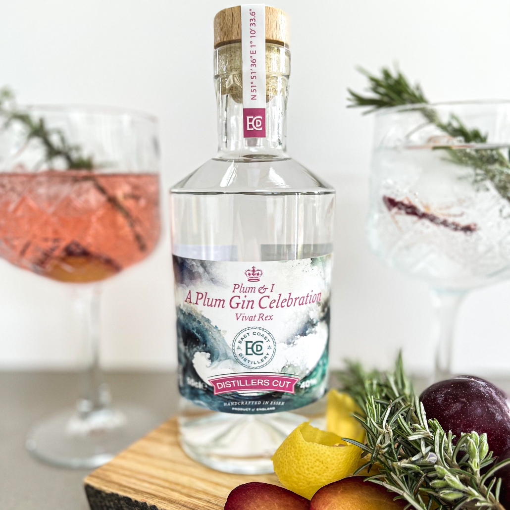 A bottle of plum gin, with two glasses out of focus in the background. Lemon peel, rosemary and plums sit on a chopping board in the foreground.